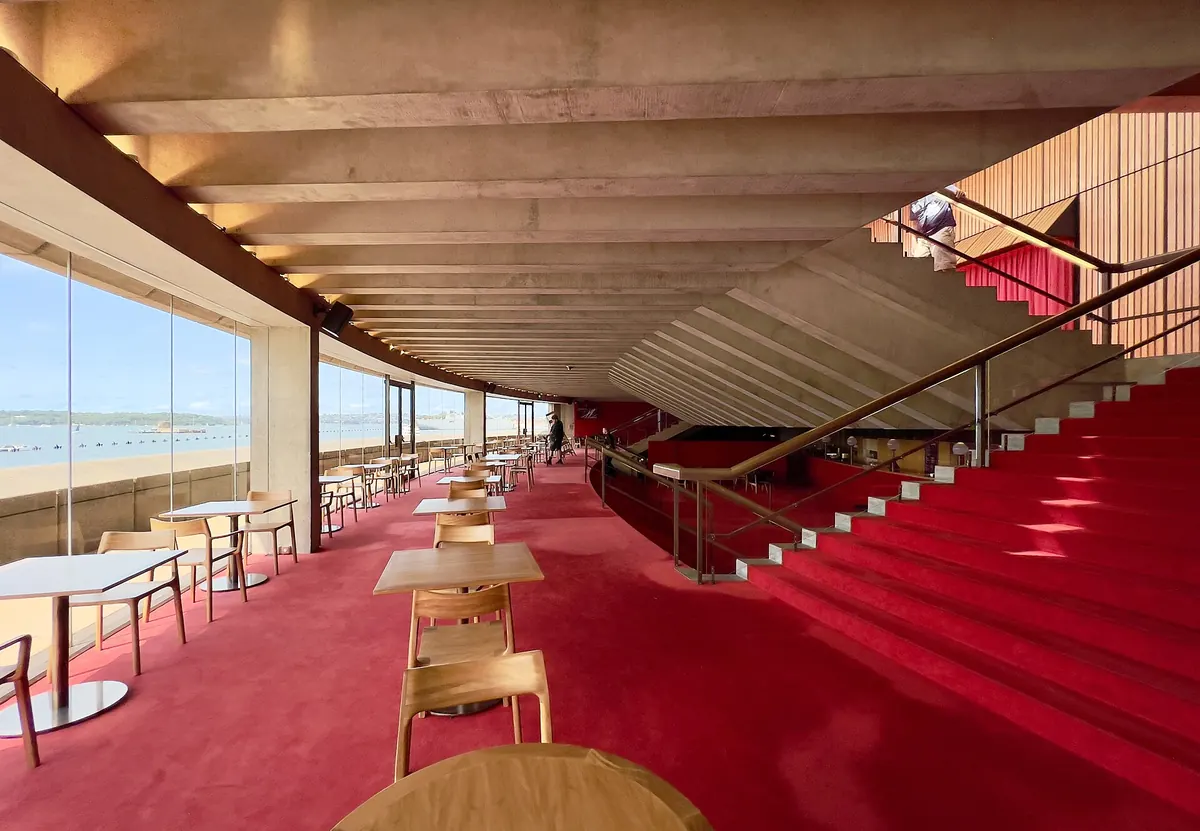 Un foyer del Joan Sutherland Theatre con moquette rossa, un soffitto in cemento a nervature e alte finestre affacciate sul porto