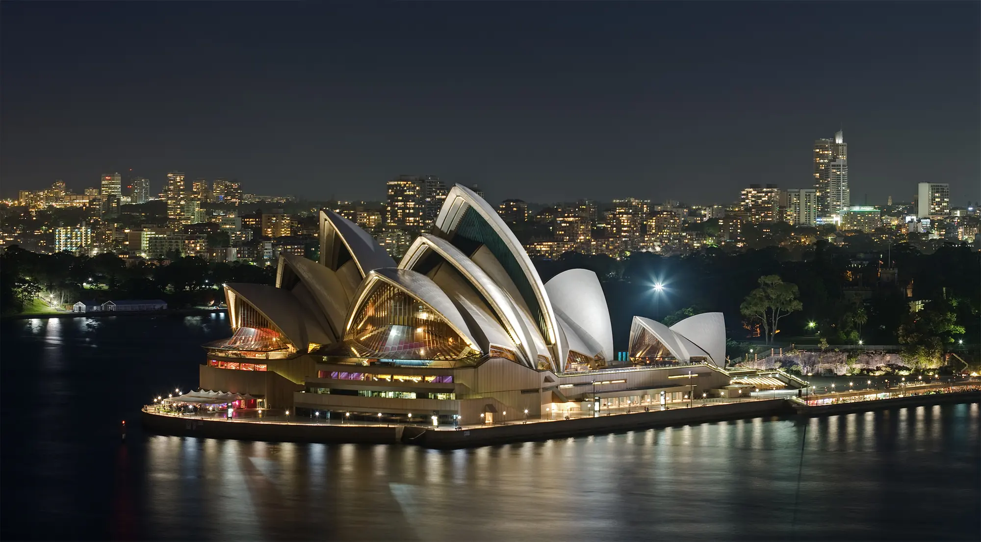 La Sydney Opera House illuminata di notte a Bennelong Point, vista dall'altra parte dell'acqua, con le luci della città sullo sfondo