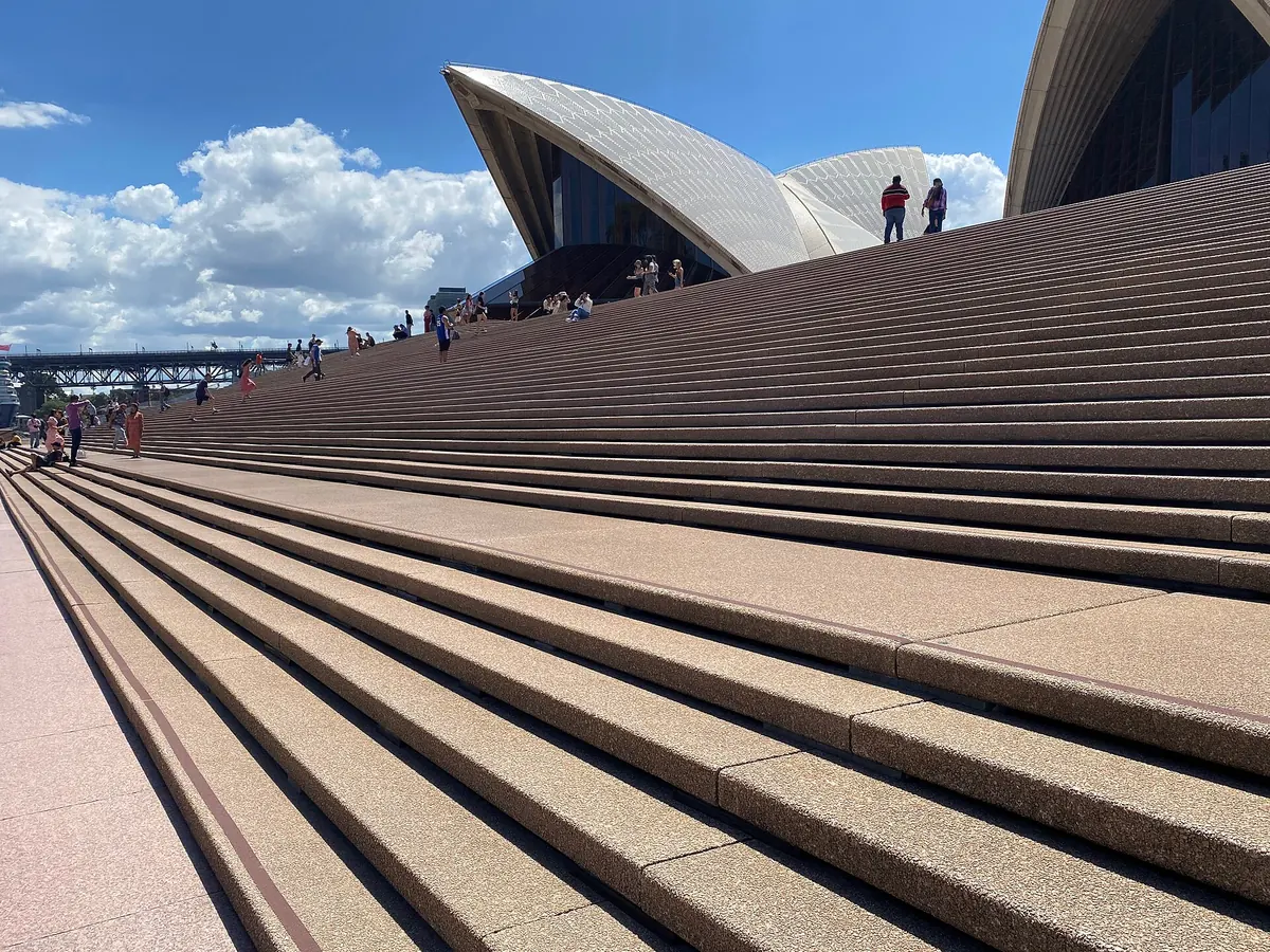 L'ampia scalinata in granito della Sydney Opera House che sale verso le conchiglie, con visitatori seduti sotto un cielo azzurro