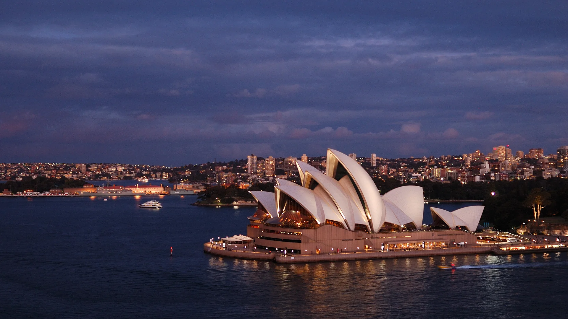 La Sydney Opera House al crepuscolo, le sue vele bianche illuminate sopra le acque scure del porto di Sydney, con le luci della città sullo sfondo